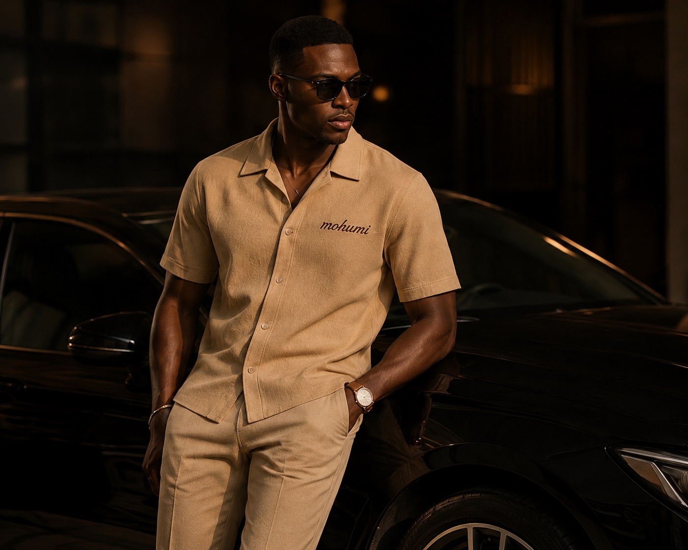 MOHUMI campaign — camel camp-collar shirt embroidered with the mohumi wordmark, worn beside a black car at night